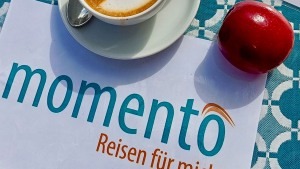 momento Reisen Logo auf einem Frühstückstisch mit einem roten Apfel und einer Tasse Kaffee